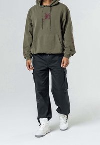 Man in een olijfgroene hoodie met rode borduursels, zwarte cargobroek en witte hoge sneakers staat tegen een effen achtergrond.