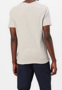 Mand iført en lysegrå t-shirt med korte ærmer og mørkeblå cargo-bukser set fra ryggen mod en hvid baggrund.
