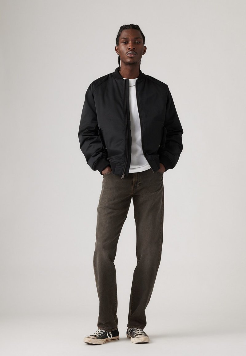 Schwarze Bomberjacke mit gerippten Bündchen, weißes T-Shirt, olivgrüne Hose und schwarze Sneaker mit weißen Sohlen. Neutraler Hintergrund.