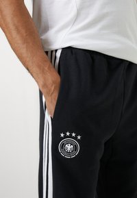 Sorte sweatpants med hvide sidelæg og et cirkulært mærke, der viser et våbenskjold med fire stjerner. Materialet ser blødt og tekstureret ud.