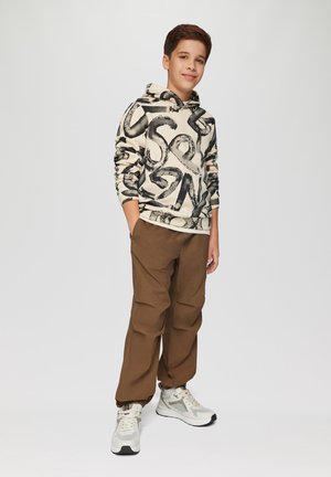Kapuzensweatshirt in Off-White mit schwarzen abstrakten Mustern, kombiniert mit braunen Hosen und hellgrauen Sneakers, die Mesh-Details aufweisen.
