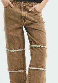 Rjave denim hlače z raztrganimi robovi pri kolenih. Imajo visok pas, ravno krojenje in žepa spredaj.