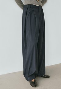 Dunkelblaue, weit geschnittene Hosen mit vorderen Falten, ausgestattet mit einem Knopfverschluss und glatter Stofftextur. Kombiniert mit schwarzen Slippers.