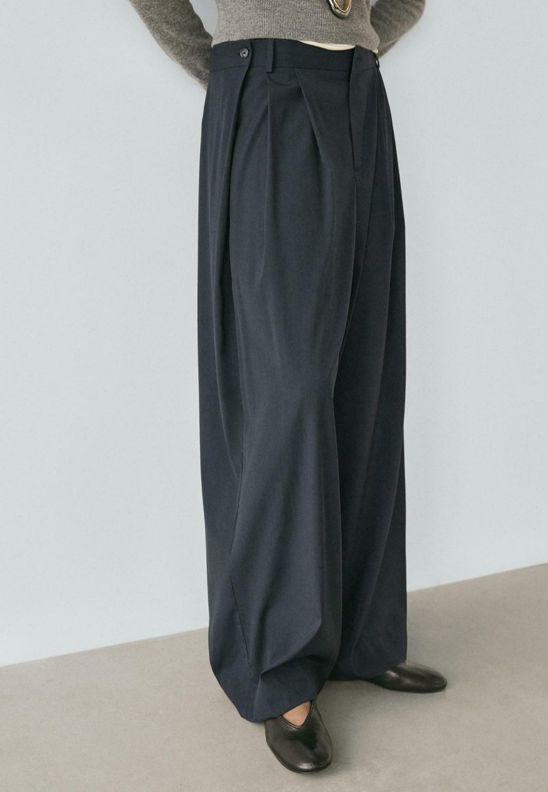 Dunkelblaue, weit geschnittene Hosen mit vorderen Falten, ausgestattet mit einem Knopfverschluss und glatter Stofftextur. Kombiniert mit schwarzen Slippers.