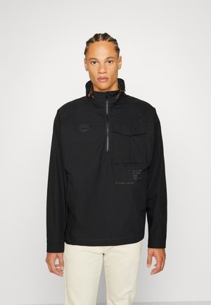 Veste noire à zip quart, avec un col montant, dotée d'une poche frontale, d'un branding subtil et de détails de couture minimalistes. Texture lisse, coupe décontractée.