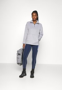 Pullover cinzento em fleece com fecho de meio zíper, combinado com leggings azul-escuro com riscas refletoras, botas pretas e uma mochila cinzenta.