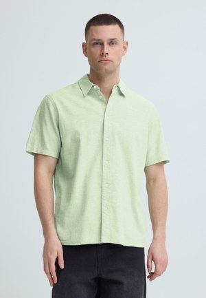 Homme aux cheveux courts portant une chemise à manches courtes vert clair boutonnée et un pantalon noir, debout devant un fond clair uni.