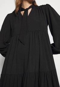 Robe noire en tissu léger, avec un nœud au cou, des manches longues et un design de jupe à volants avec une coupe ample.