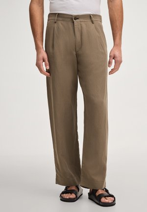 Mężczyzna noszący luźne khaki spodnie i czarne sandały z klamrą, stojący na tle jasnego, jednolitego tła.