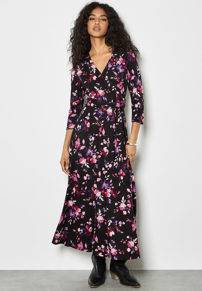 Vestido negro de manga larga con estampado floral en tonos rosas y morados. Presenta un escote en V, cintura con lazo y falda fluida. Se lleva con botas negras.