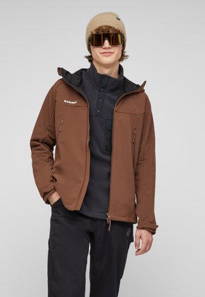 Giovane uomo che indossa una giacca Mammut marrone, pantaloni neri, berretto beige e grandi occhiali da sole riflettenti, sorridente con le mani nelle tasche.
