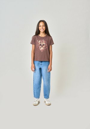 Jeune fille aux longs cheveux portant un t-shirt marron avec un motif de crâne floral, un jean bleu et des baskets blanches, debout et souriante.
