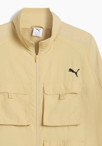 Ljusgul jacka med hög krage, som har två framfickor och en svart Puma-logotyp på vänster bröstområde.