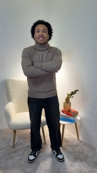 Pull beige à col roulé, jean foncé, baskets noires avec des accents blancs. Debout à côté d'un fauteuil crème et d'une petite table avec des livres.