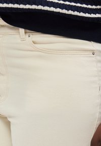 Κοντινό ενός ατόμου που φοράει κρεμ παντελόνι και μπλε σκούρο πουλόβερ με λευκές ρίγες στο τελείωμα.