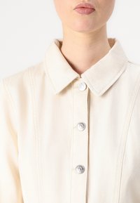 Veste en denim crème avec un col pointu, des boutons-pression en métal argenté et des détails de couture verticale. Le tissu semble durable avec une texture douce.