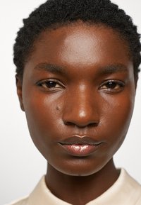 Close-up van een zwarte persoon met kort krullend haar, een egale huid, glanzende lippen en een neutrale uitdrukking, gekleed in een lichtgekleurd shirt.