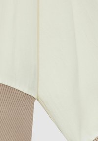 Krämfärgad topp med en mjuk textur, med en ribbad beige nederkant. Designen inkluderar en avslappnad passform och lager element.