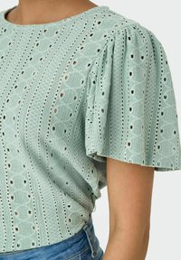 Hellgrüne, strukturierte Bluse mit gepufften Kurzärmeln und Lochmuster. Runde Ausschnittform und lockere Passform. Sichtbare Jeans-Shorts.