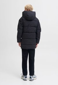 Melna pūkaina jaka ar kapuci, ar izšūtu rakstu un vidēja garuma griezumu. Valdīta virs melnām biksēm un baltām sporta kurpēm.