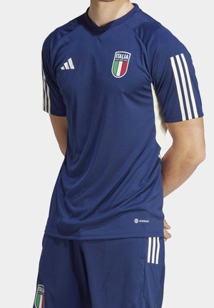 Homme portant un maillot de football bleu de l'Italie et un short avec des rayures blanches et le blason de l'Italie sur la poitrine et la jambe, debout les mains derrière le dos.