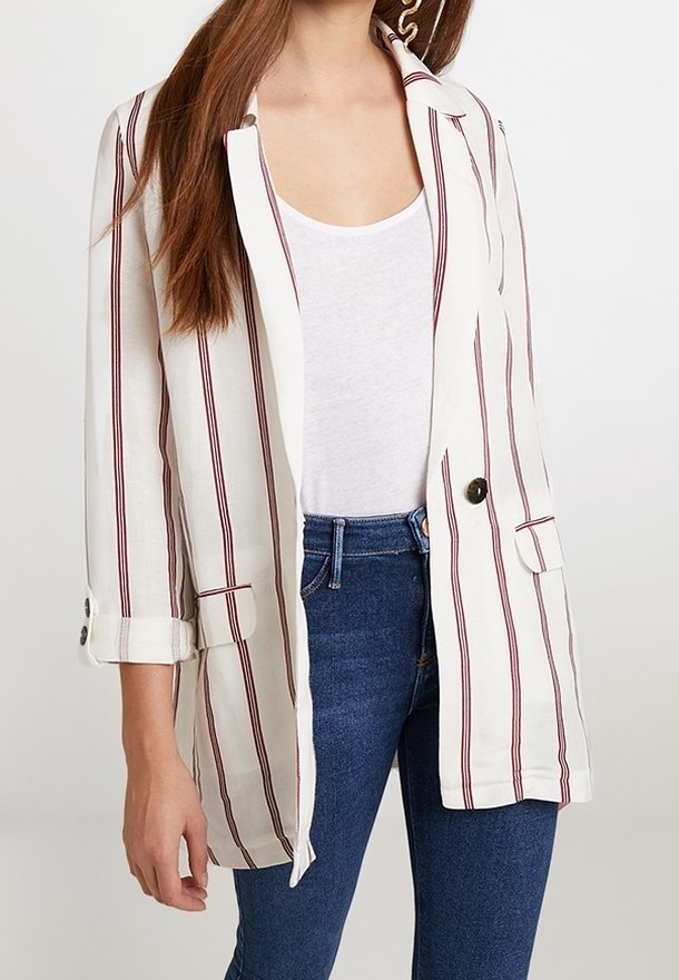 Femme portant un blazer blanc à rayures rouges verticales, un débardeur blanc et un jean bleu, debout devant un fond uni.