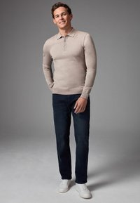 Beige Langarm-Polohemd mit drei Knöpfen, kombiniert mit dunkelblauen Jeans und weißen Sneakern. Glatte Textur und taillierte Passform.