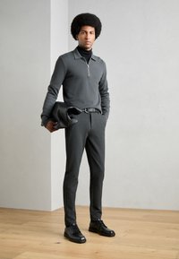 Temno siva pulover z zadrgo, črn pulover s turtleneck ovratnikom, krojeni hlače, črn pas in čevlji, drži črn clutch, stoji na lesenem tlu.
