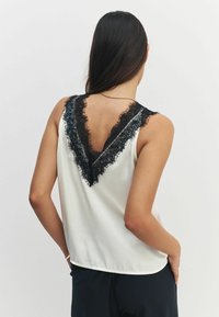 Bez piedurknēm, baltas satīna blūze ar dziļu V formas aizmuguri, ar sarežģītu melnu mežģīņu apdari ap dekoltē un aizmugurējām malām.