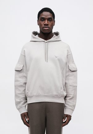 Uomo che indossa una felpa con cappuccio grigio chiaro con tasche a soffietto sulle maniche e pantaloni scuri, in piedi su uno sfondo bianco semplice.