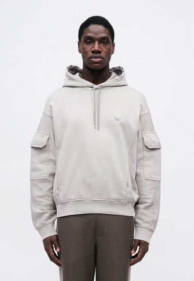 Uomo che indossa una felpa con cappuccio grigio chiaro con tasche a soffietto sulle maniche e pantaloni scuri, in piedi su uno sfondo bianco semplice.
