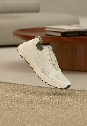Witte lichtgewicht sneaker met gebreid bovenwerk en dikke zool, zwevend boven een beige tapijt nabij een houten ronde salontafel met boeken.