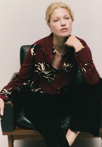 Cămașă burgundy cu guler, cu un model floral în nuanțe de crem, asortată cu pantaloni negri. Modelul este așezat pe un scaun negru tapițat. Accente de bijuterii aurii.