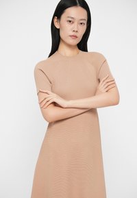 Vestido de malha bege com mangas curtas e textura canelada, apresentando um corpete justo e uma saia fluida, acentuando uma silhueta elegante.