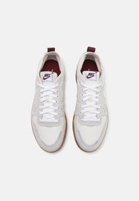 Sneaker Nike bianche con tomaia in tessuto liscio, tocchi di bordeaux sul tallone e suola in gomma naturale con un motivo testurizzato.