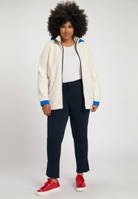 Chaqueta de abrigo blanca con cremalleras, con detalles en azul, combinada con pantalones marinos con cordón. El modelo lleva zapatillas rojas. Diseño simple y casual.