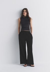 Haut noir sans manches avec col montant, associé à un pantalon large noir et ample. Les matériaux semblent doux avec une texture lisse.