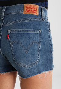 Farkkushortsit haalistuneessa sinisessä värissä, joissa on kuluneet helmat, ruskea nahkapaikka, jossa lukee "Levi Strauss & Co.", ja kaarevat takataskujen saumat.