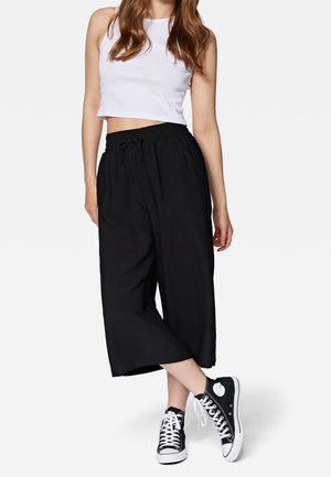 Vrouw draagt een wit mouwloos cropped topje, zwarte wijde cropped broek en zwarte hoge sneakers met witte veters op een witte achtergrond.