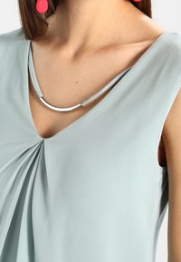 Kvinna bär en ljusblå ärmlös topp med veckdetalj fram, silverfärgad böjd halskedja och röda hängande örhängen.