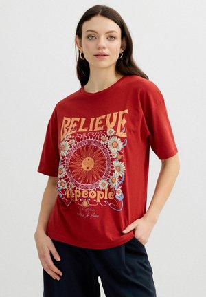 Joven mujer con cabello largo y oscuro, lleva pendientes de aro dorados y una camiseta roja con diseño gráfico de sol y flores, de pie con la mano en el bolsillo.