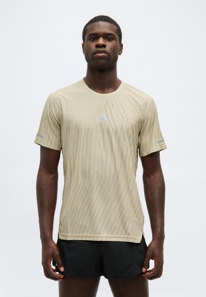 Mand iført beige tekstureret kortærmet træningstrøje og sorte shorts, stående mod en ensartet lys grå baggrund.