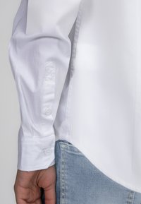 Witte button-up shirt met een schone, gladde textuur, een afgeronde zoom en knoopmanchetten, gecombineerd met lichtblauwe denim.
