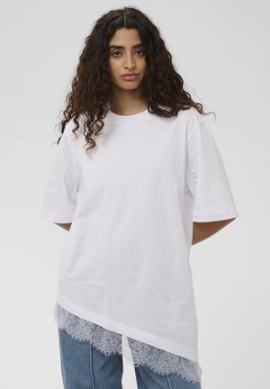 Noor naine lokkis tumedate juustega, seljas lahtine valge pitsilise äärega T-särk ja sinised teksad, seisab ühetaustal helerohelise tausta ees.