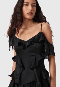 Donna con lunghi capelli ricci che indossa un vestito nero senza spalline con volant sovrapposti e sottili spalline trasversali, su uno sfondo semplice.