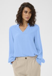 Blouse bleu clair à col en V, manches longues avec poignets froncés, assortie à un pantalon beige. Texture de tissu lisse et léger.