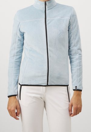 Personne portant une veste en polaire bleu clair avec une fermeture éclair noire et des finitions noires, assortie à un pantalon blanc avec des poches zippées noires.