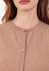 Femme portant un cardigan beige en maille avec deux boutons ronds près du col et de la poitrine, montrant une partie de ses lèvres et de son cou.