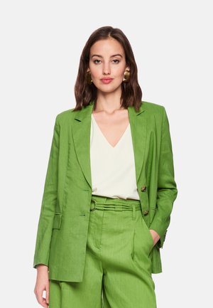 Junge Frau trägt einen leuchtend grünen Anzug mit weißer Bluse, goldene auffällige Ohrringe, posiert mit einer Hand in der Tasche vor einem schlichten Hintergrund.