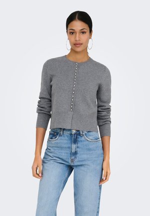 Junge Frau mit dunklem Haar in einem grauen Pullover mit Knöpfen und hellblauen hoch taillierten Jeans, stehend vor einem schlichten hellen Hintergrund.